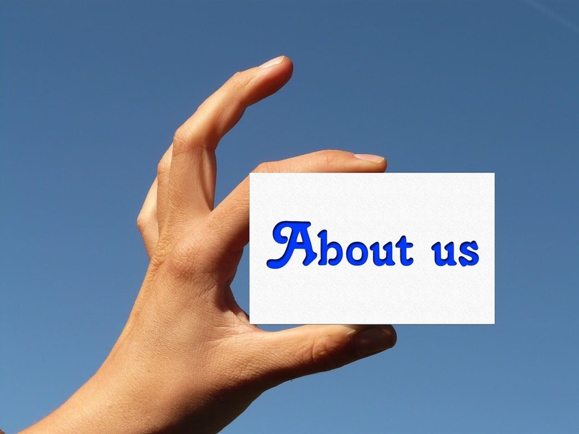 About Us 1 A hand holding a white card with the blue text “About us” against a clear blue sky background, representing a website’s About Us page.