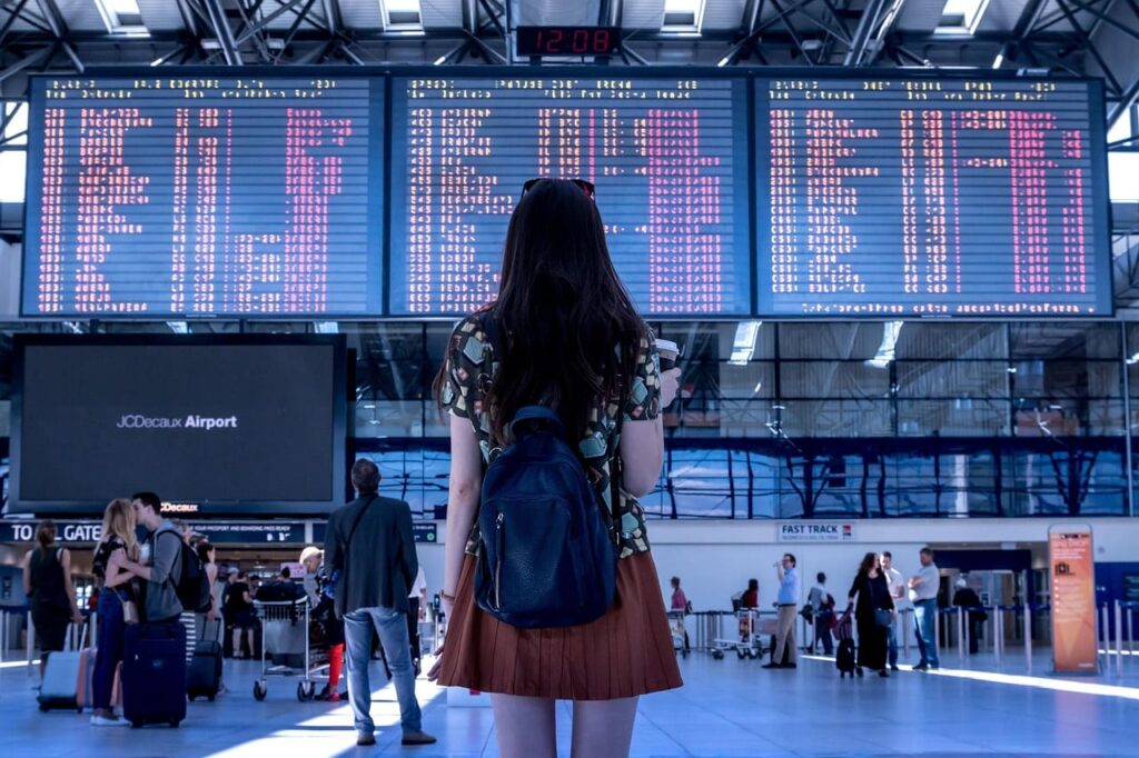 U.S. to Require Visa Bonds for Tourists: What Travelers Need to Know 2 A woman with long hair and a backpack stands in an airport, facing a large electronic departure board full of flight information.